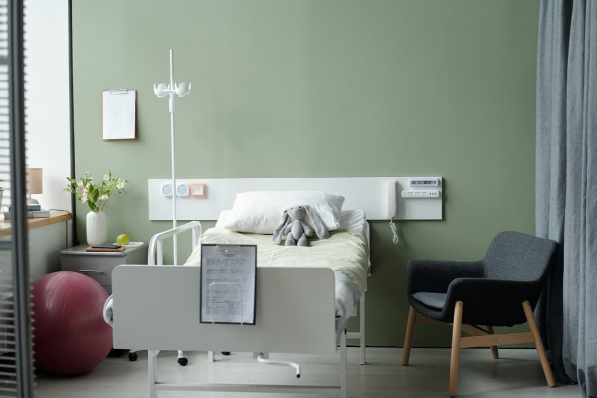 Nemocniční pokoj s postelí, křeslem a zelenou stěnou.